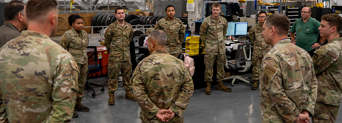 A group of uniformed U.S. Air Force personnel stand in a circle inside a maintenance facility surrounded by equipment and tires.